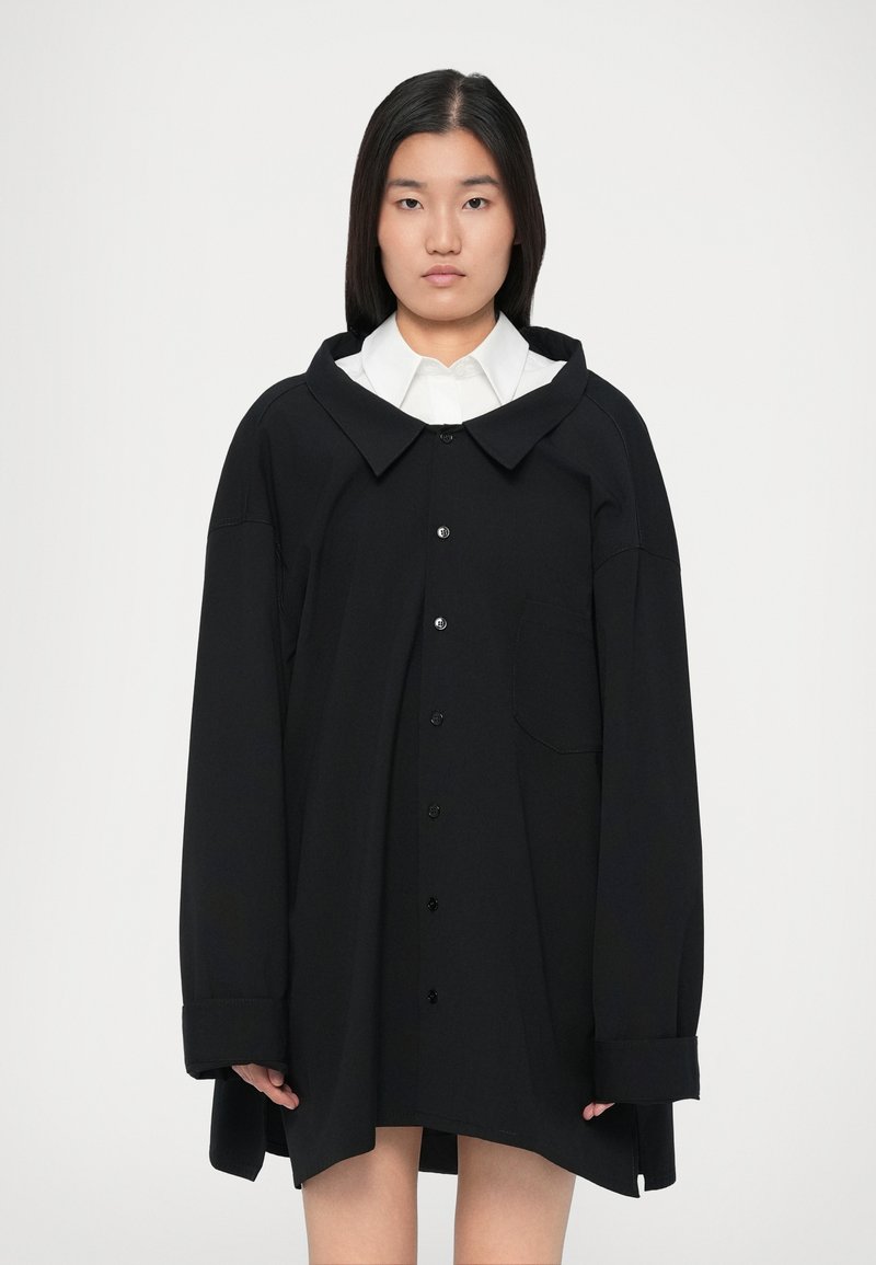 Ung kvinde med s Straight mørkt hår iført en oversized sort knapjakke over en hvid skjorte, stående mod en ensfarvet baggrund.