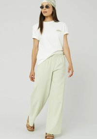 T-shirt en coton blanc avec des manches roulées, pantalons amples à carreaux verts, et sandales à plateforme marron. Lunettes de soleil et foulard à motifs inclus.