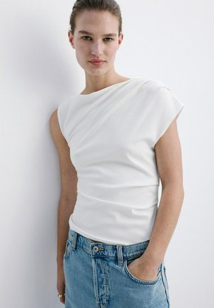 Femeie cu părul prins la spate, purtând un top alb fără mâneci și blugi albaștri, sprijinită de un perete alb cu o mână în buzunar.