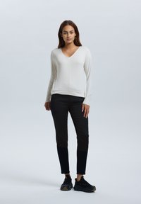 Lichtbeige langemouwen top met een V-hals, gecombineerd met zwarte slim-fit broeken met gestructureerde zwarte accenten, en zwarte sneakers.