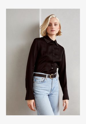 Melna krekla ar apkakli, apvienota ar gaiši ziliem džinsiem un melnu jostu ar zelta apaļu sprādzi.