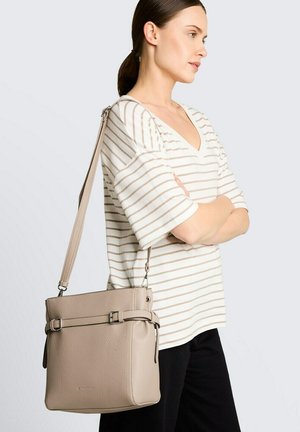 Frau mit braunen Haaren in einem Pferdeschwanz, die ein weiß gestreiftes V-Ausschnitt-Shirt und schwarze Hosen trägt, mit einer beigen Leder-Umhängetasche.