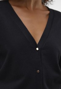 Cardigan negru cu decolteu în V, realizat dintr-un material tricotat, având accente subtile cu nasturi aurii în partea din față. Textură netedă și design potrivit.