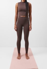 Brun sportig crop top och högmidjade leggingar, tillverkade av glatt, elastiskt material. Stående barfota på en rosa yogamatta.