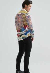 Homme en pantalon et chaussures noires portant une chemise à manches longues colorée avec des motifs de soleil et d'éléments célestes, se tenant de côté devant un fond uni.