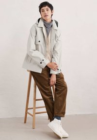 Vaaleanharmaa takki, jossa on vihreä huppu, yhdistettynä raidalliseen poolopaitaan ja vaaleanbeigeen neuleeseen, ruskeisiin corduroy-housuihin ja valkoisiin lenkkareihin.