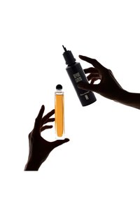 Bottiglia di profumo in vetro con tappo sferico nero, riempita di liquido ambrato; flacone spray nero con testo bianco sorretto da mani, su uno sfondo chiaro.