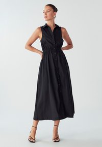 Juoda, be rankovių maxi suknelė su mygtukų uždarymu priekyje ir elastinga juosmeniu. Lygus audinys su klasikine apykakle ir minimalistiniu dizainu.