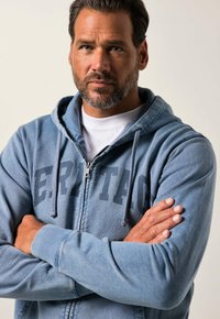 Hooded zip-up sweatshirt in light blue fabric, featuring a front logo, ribbed cuffs, and drawstrings. Model stands with arms crossed.