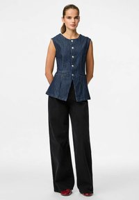 Chaleco de denim a rayas azules con cuerpo ajustado, escote en forma de scoop, seis botones frontales y dobladillo acampanado. Combinado con pantalones anchos negros y zapatos rojos.