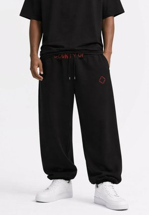 Mand iført sorte løse sweatpants med rødt detaljer og hvide sneakers, hænderne i lommerne, stående mod en hvid baggrund.