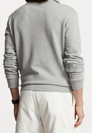 Persoon draagt een lichtgrijze lange mouwen sweatshirt met gerimpelde mouwen en een witte broek, gezien van achteren tegen een effen achtergrond.