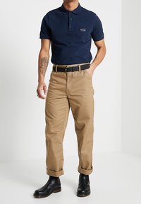 Marinblå polo skjorta med tre knappar och en etikett, tillsammans med beige chinos som är uppvikta vid fållen och svarta ankelstövlar.