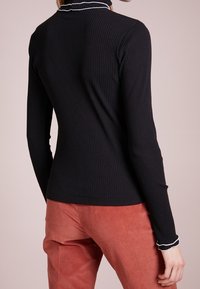 Haut noir côtelé à manches longues avec un biais blanc au col, associé à un pantalon en velours côtelé de couleur rouille. Texture lisse, coupe ajustée.