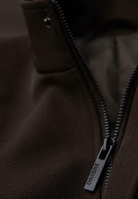 Chaqueta marrón con cremallera y un tejido texturizado. Presenta una cremallera negra resistente y una pequeña etiqueta que dice "OMBRE" cerca de la cremallera.