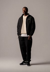 Črna bomber jakna čez kremast pulover iz pletenine, kombinirana s črnimi športnimi hlačami in črnimi supergami z belimi poudarki. Preprosta, priložnostna obleka.