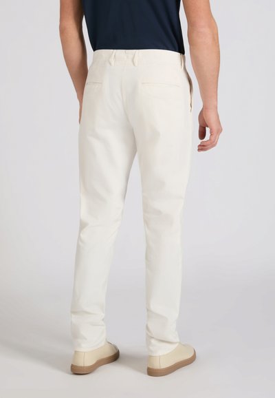 Hombre con pantalones rectos color blanco roto, zapatos beige y una camisa oscura metida, de pie de espaldas sobre un fondo liso.