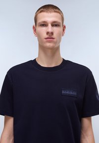 Camiseta de algodón azul marino con cuello redondo, con un pequeño logo en parche en el lado izquierdo del pecho. Textura suave y corte casual.