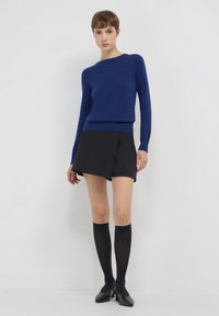 Marineblauer Strickpullover mit langen Ärmeln, kombiniert mit einem schwarzen Wickel-Minirock und knielangen schwarzen Socken, abgerundet mit schwarzen Schuhen.