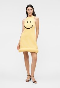Femme portant une mini-robe jaune sans manches avec un visage souriant noir, associée à des talons noirs à bride à la cheville, debout devant un fond uni.