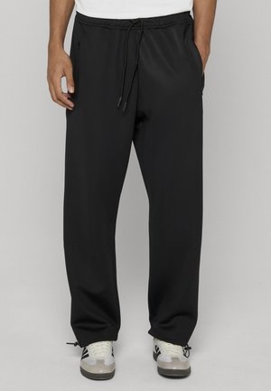 Mężczyzna noszący czarne, szerokie spodnie z troczkiem w pasie i zapinanymi na zamek bocznymi kieszeniami, w połączeniu z białymi i czarnymi trampkami.