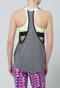 Top grigio senza maniche con schiena aperta, caratterizzato da un design a strati e dettagli neri. Abbinato a leggings rosa a fantasia che mostrano forme geometriche.