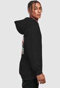 Sudadera negra con bolsillo canguro en la parte frontal, con un gráfico colorido de flores y texto en el lado izquierdo. Tejido suave, corte regular.