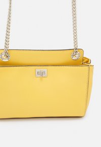 Borsa in pelle gialla con forma strutturata. Presenta una tracolla a catena argentata, dettaglio con logo e superficie texturizzata.