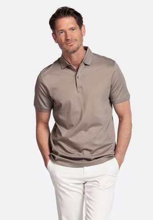 Man staat met handen in zakken, draagt een taupekleurig poloshirt met korte mouwen en witte broek tegen een effen achtergrond.