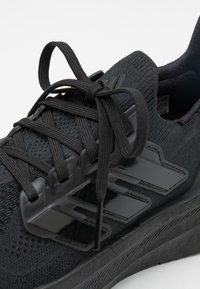 Chaussure de sport noire avec une tige en tissu texturé, des lacets noirs et des accents noirs brillants. La semelle présente un motif de traction distinct pour une adhérence optimale.