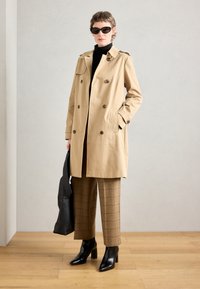 Beige dubbelknäppt trenchcoat med axelklaffar, kombinerad med bruna rutiga byxor och svarta klackboots. Accessoarer med en svart väska.