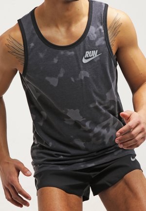 Mężczyzna ubrany w ciemną kamuflażową koszulkę bez rękawów z logo "RUN" i charakterystycznym znakiem swoosh oraz czarne krótkie spodenki do biegania, w pozie biegacza z tatuażami na górnych partiach ramion.