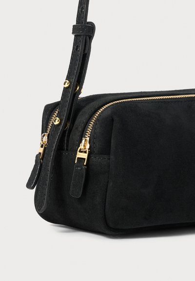 Borsa rettangolare nera in suede con cerniere dorate e tracolla regolabile con borchie dorate su sfondo bianco.