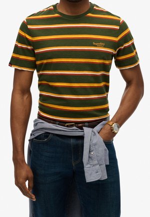 Mężczyzna w zielono-pomarańczowo-białej koszulce w paski z logo "SuperDry Athletics", ciemnych dżinsach, zegarku i szarej koszuli związanej wokół talii.