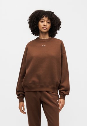 Žena s kučeravými vlasmi v hnedej mikine Nike a zodpovedajúcich hnedých teplákoch Nike stojaca pred jednofarebným svetlým pozadím.