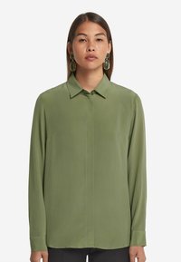 Grünes Langarmshirt mit Kragen, aus glattem Stoff gefertigt. Verfügt über eine durchgehende Knopfleiste und einen geraden Saum. Ein Paar kräftige grüne Ohrringe.