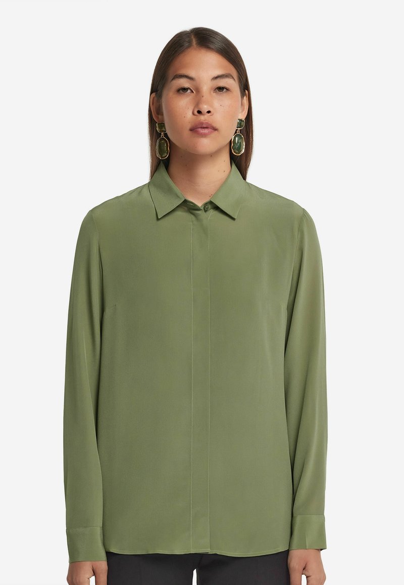 Grünes Langarmshirt mit Kragen, aus glattem Stoff gefertigt. Verfügt über eine durchgehende Knopfleiste und einen geraden Saum. Ein Paar kräftige grüne Ohrringe.