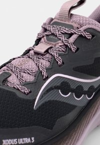 Zapatilla de trail running negra y gris con una parte superior de malla texturizada, cordones morados, un logotipo prominente y una suela resistente para tracción.