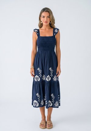 Woman wearing a navy blue sleeveless midi dress with white floral embroidery, ruffled shoulder straps, and tan sandals, standing against a plain background.