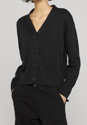 Persoon draagt een zwarte, met knopen gesloten V-hals vest met lange mouwen en zwarte broek, handen in de zakken, tegen een effen achtergrond.