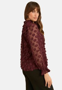 Kvinna som bär en genomskinlig bordeauxblus med upphöjda prickar och volangärmar, tillsammans med svarta byxor, vänd lite på sidan.