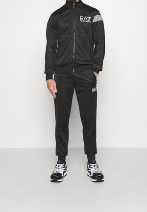 Mann trägt schwarzen EA7 Emporio Armani Trainingsanzug mit weißen Logodetails und schwarz-weißen Sneakers, steht vor einfarbigem Hintergrund.