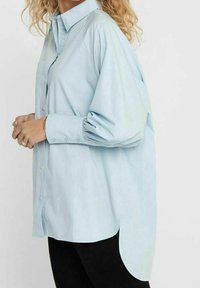Camicia azzurra chiaro con bottoni, dal taglio morbido, maniche a sbuffo e orlo curvato. Realizzata in tessuto morbido con una consistenza liscia.