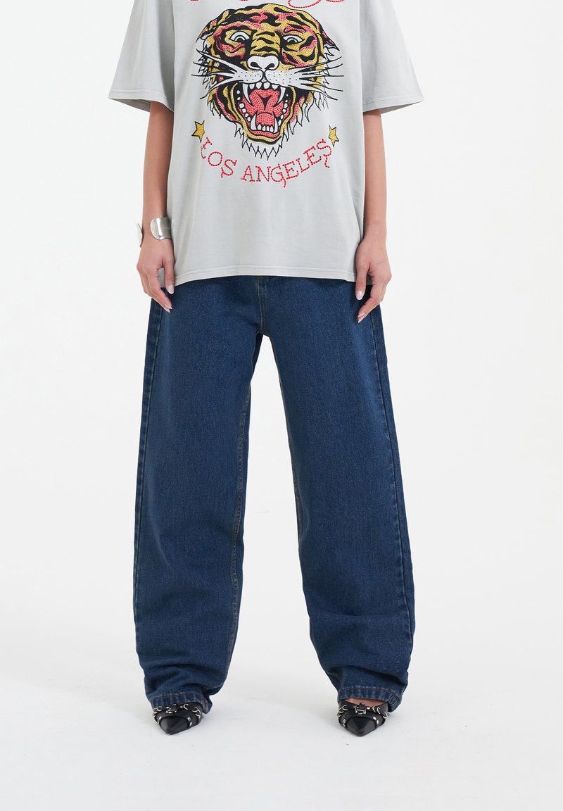 Grijze oversized t-shirt met een kleurrijke tijgerafbeelding, gecombineerd met losse blauwe denim jeans en zwarte puntige schoenen. Eenvoudige armbandaccessoire.