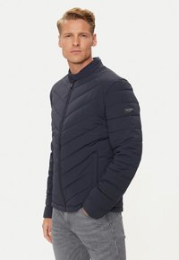 Giacca a vento blu navy con colletto alto, motivo a rombi, chiusura con zip e patch con logo sul maniche sinistro. Finitura testurizzata.