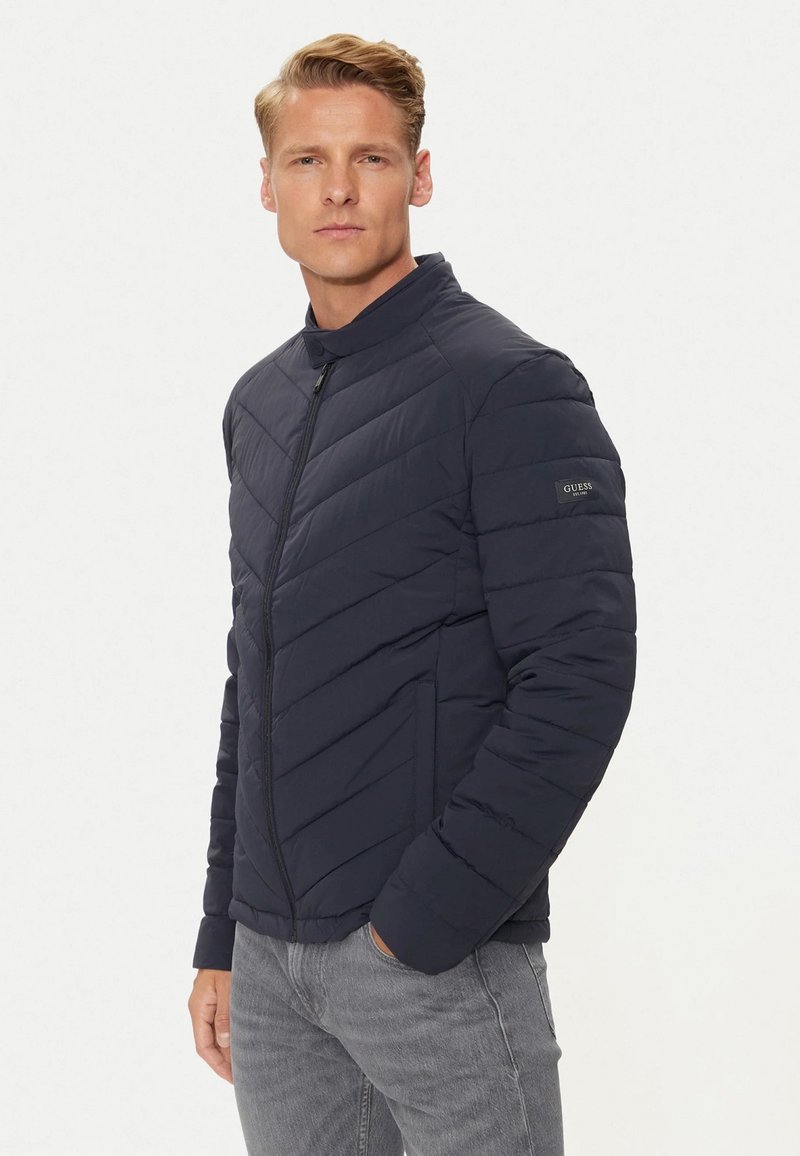 Giacca a vento blu navy con colletto alto, motivo a rombi, chiusura con zip e patch con logo sul maniche sinistro. Finitura testurizzata.