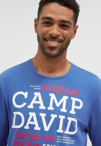 Lächelnder Mann mit kurzen lockigen Haaren, der ein blaues Hemd mit weißem und rotem "CAMP DAVID"-Schriftzug vor einem schlichten hellen Hintergrund trägt.