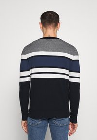 En man som står med ryggen mot kameran, iklädd en randig tröja i marinblått, vitt och grått samt blå jeans, framför en neutral bakgrund.