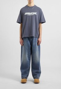 Szara koszulka z dużym napisem "AVIREX DEPT.", zestawiona z luźnymi jasnoniebieskimi dżinsami z wybledłymi akcentami oraz jasnobrązowym obuwiem.