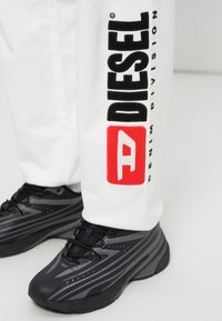 Λευκά παντελόνια jogging με την επιγραφή «DIESEL» και κόκκινο λογότυπο στο αριστερό πόδι, σε συνδυασμό με μαύρα αθλητικά παπούτσια με γκρι λεπτομέρειες και υφή στις σόλες.